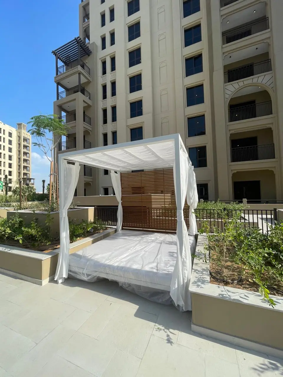 Close-up of a custom pool cabana with matte white powder-coated frame, Grade A teak slatted back panel, sheer curtains and integrated dual daybed with Sunbrella cushions